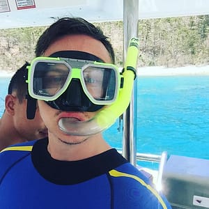 Man in SCUBA Mask and Snorkel stands in front of blue water.